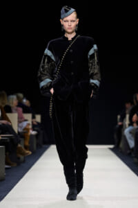 Model walking a fashion show wearing a black velvet suit with embellished patterned sleeves, a beaded diagonal accessory, and a matching hat; hands in pockets as she strides down the runway.