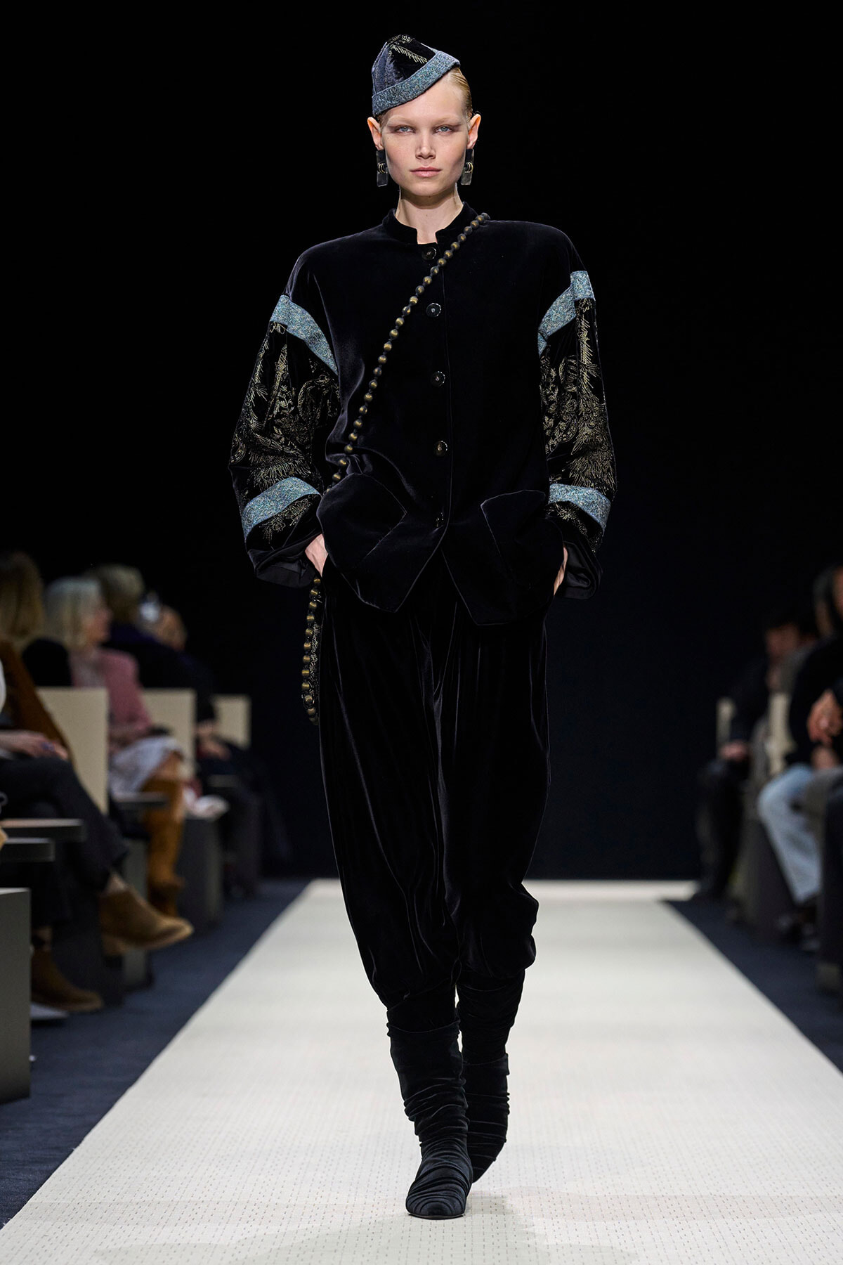 Model walking a fashion show wearing a black velvet suit with embellished patterned sleeves, a beaded diagonal accessory, and a matching hat; hands in pockets as she strides down the runway.