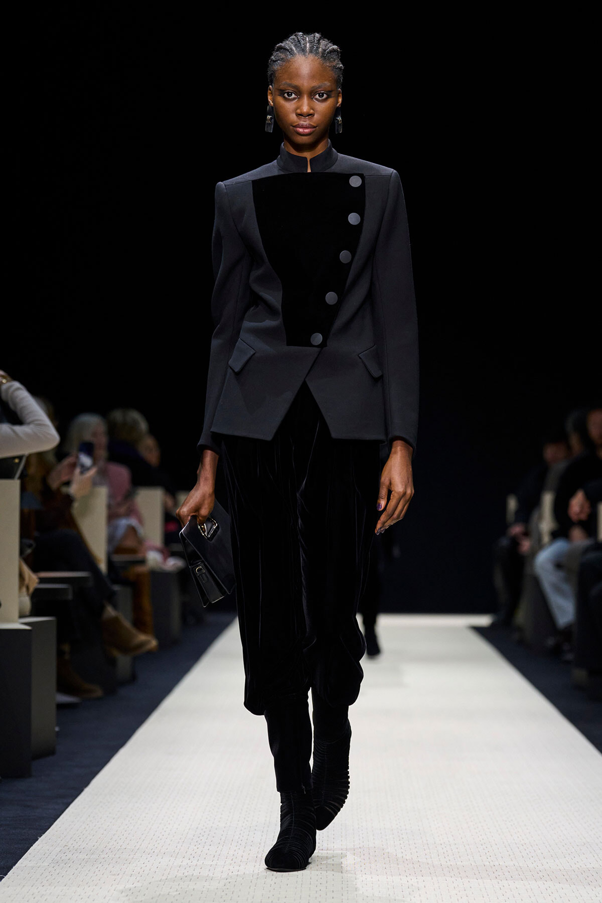 Model walking a runway in a dark, tailored black outfit with a velvet panel and large buttons, paired with matching pants and boots, holding a clutch.