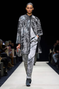 Runway model wearing a silver-gray fur coat over a marbled silver suit, holding a small metallic clutch.
