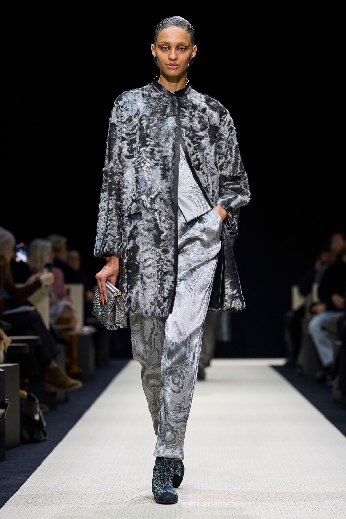 Runway model wearing a silver-gray fur coat over a marbled silver suit, holding a small metallic clutch.
