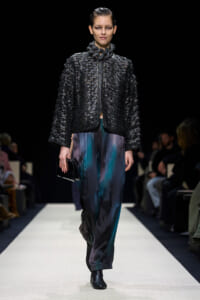 Model walking a runway in a black textured quilted jacket and teal-black striped pants, holding a small black clutch.