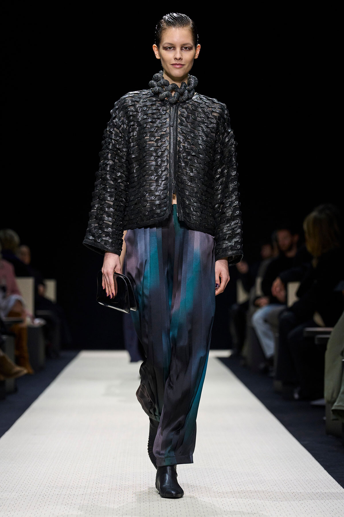 Model walking a runway in a black textured quilted jacket and teal-black striped pants, holding a small black clutch.