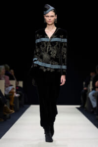 Model walking the runway in a black velvet embroidered outfit with blue bands and matching hat.
