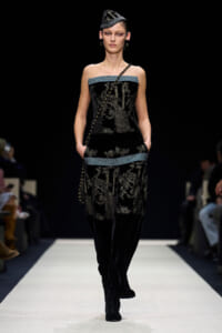 Model on a fashion runway wearing a black velvet strapless top and matching wide-leg pants with silver‑gold embroidery and blue trim; a long beaded diagonal strap and a small sculpted hat complete the look.