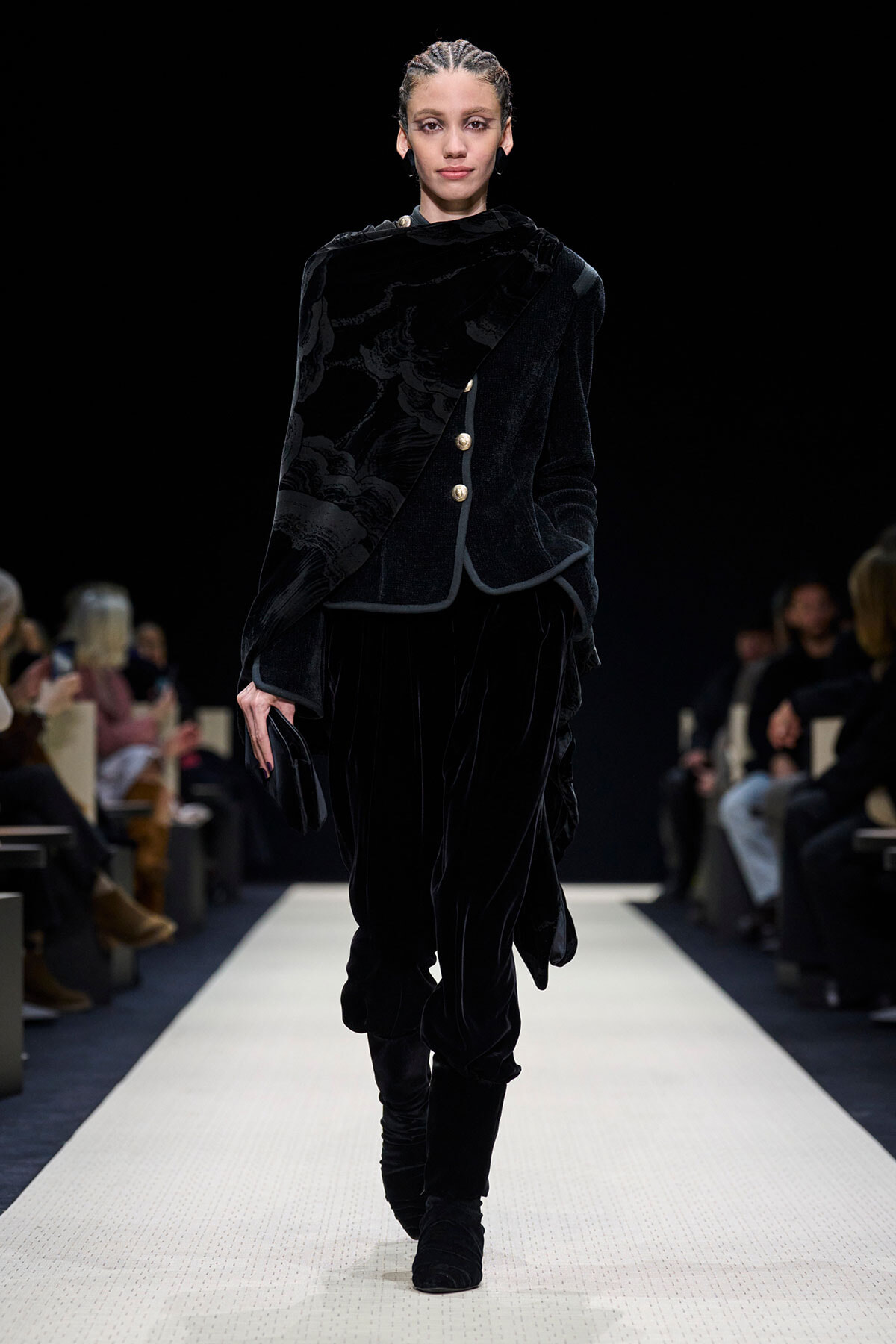 Model walks the runway in a black velvet suit with a patterned velvet scarf and gold buttons.