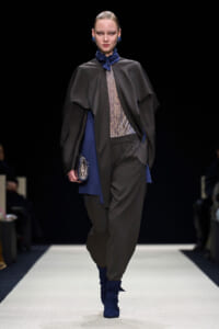 Model walks a runway wearing a dark cape over a sheer patterned top, loose black pants, and blue accessories including a scarf, boots, and clutch against a black background.