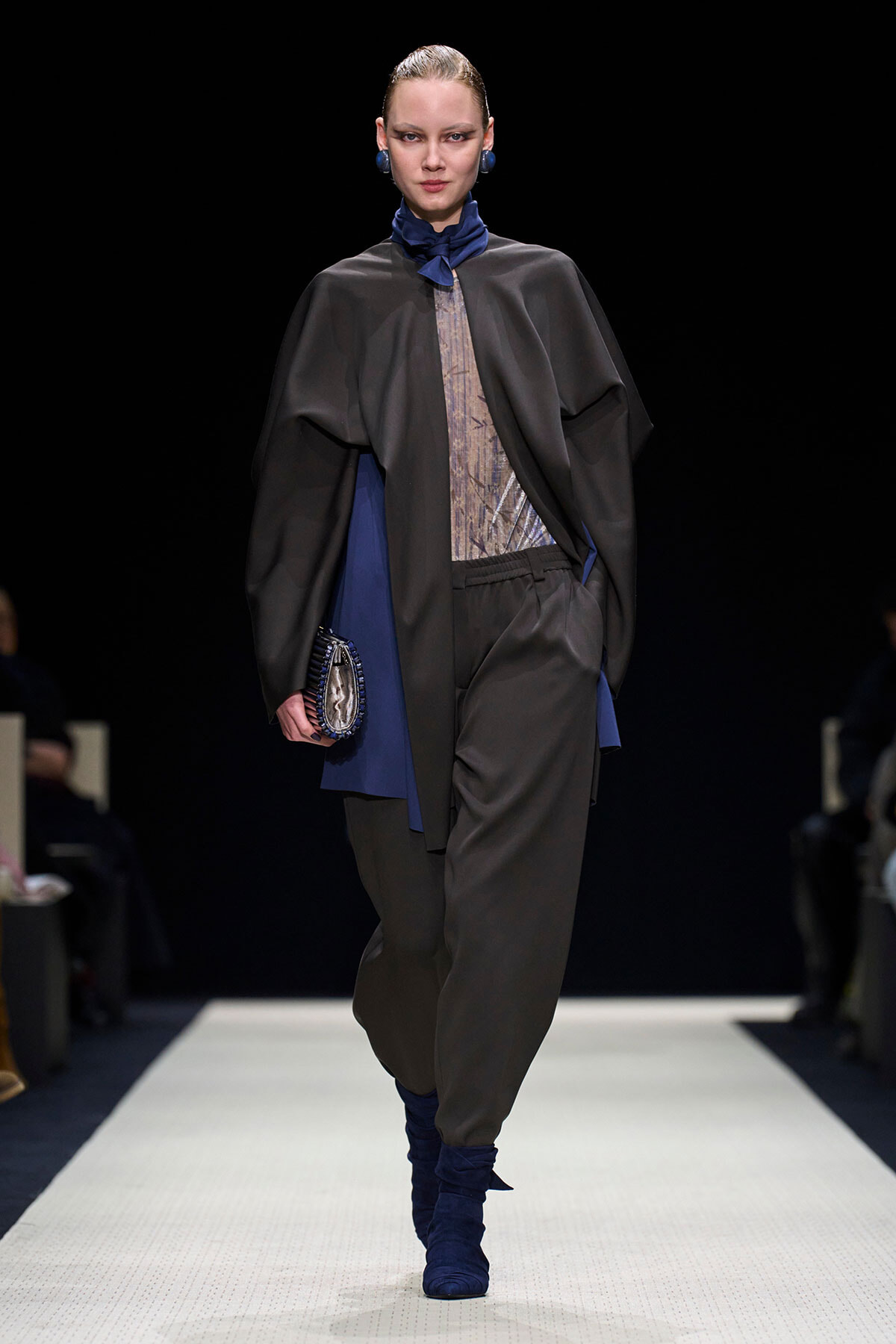 Model walks a runway wearing a dark cape over a sheer patterned top, loose black pants, and blue accessories including a scarf, boots, and clutch against a black background.