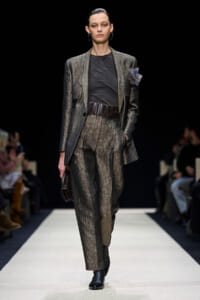 Female model walks a runway wearing a textured gray-brown suit with a black top and wide belt, pairing ankle boots.