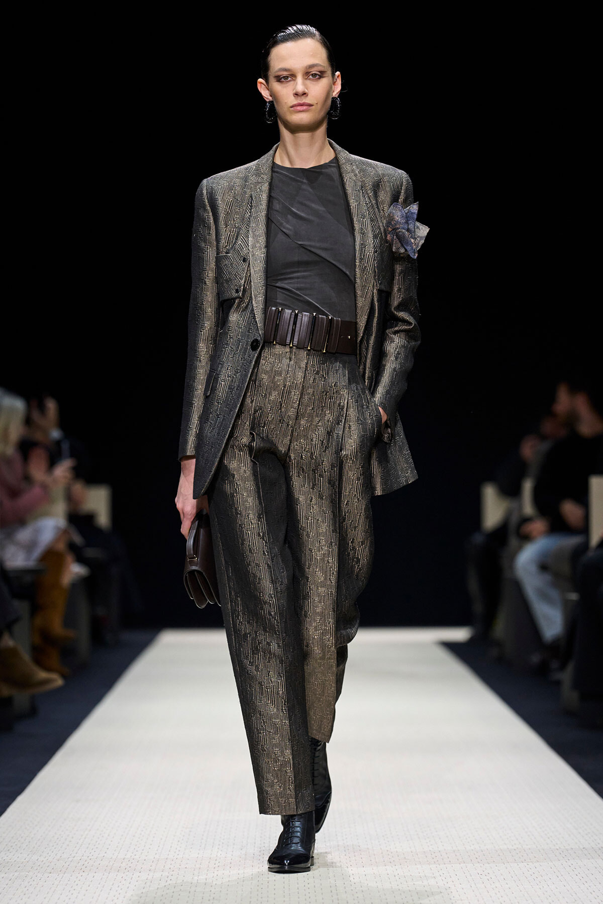 Female model walks a runway wearing a textured gray-brown suit with a black top and wide belt, pairing ankle boots.