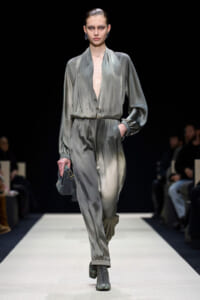 Model walks a fashion runway wearing a grey satin jumpsuit with a deep V neckline, cinched waist, and relaxed pants; she holds a small grey handbag and wears lace-up boots against a dark background.