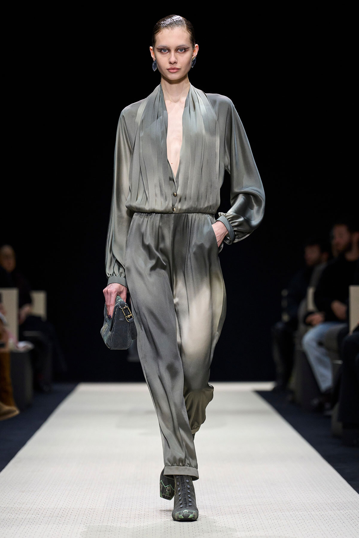 Model walks a fashion runway wearing a grey satin jumpsuit with a deep V neckline, cinched waist, and relaxed pants; she holds a small grey handbag and wears lace-up boots against a dark background.