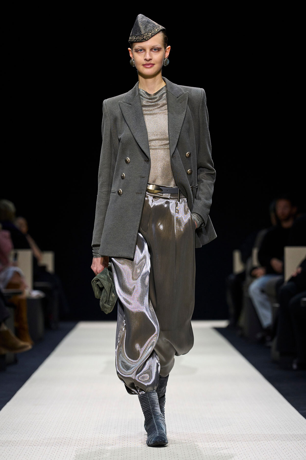 Model on a runway wearing a gray double-breasted blazer with metallic silver pants and a small patterned cap, against a black backdrop.