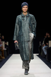 Female model walking a runway in a teal-dotted patterned coat over gray pants, wearing a dark blue headscarf and teal high-top boots, carrying a small clutch