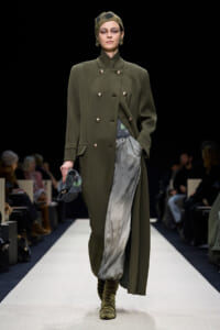 Model walking a runway in an olive double-breasted coat, metallic gray pleated pants, olive boots, and a patterned headscarf with a small clutch in hand.