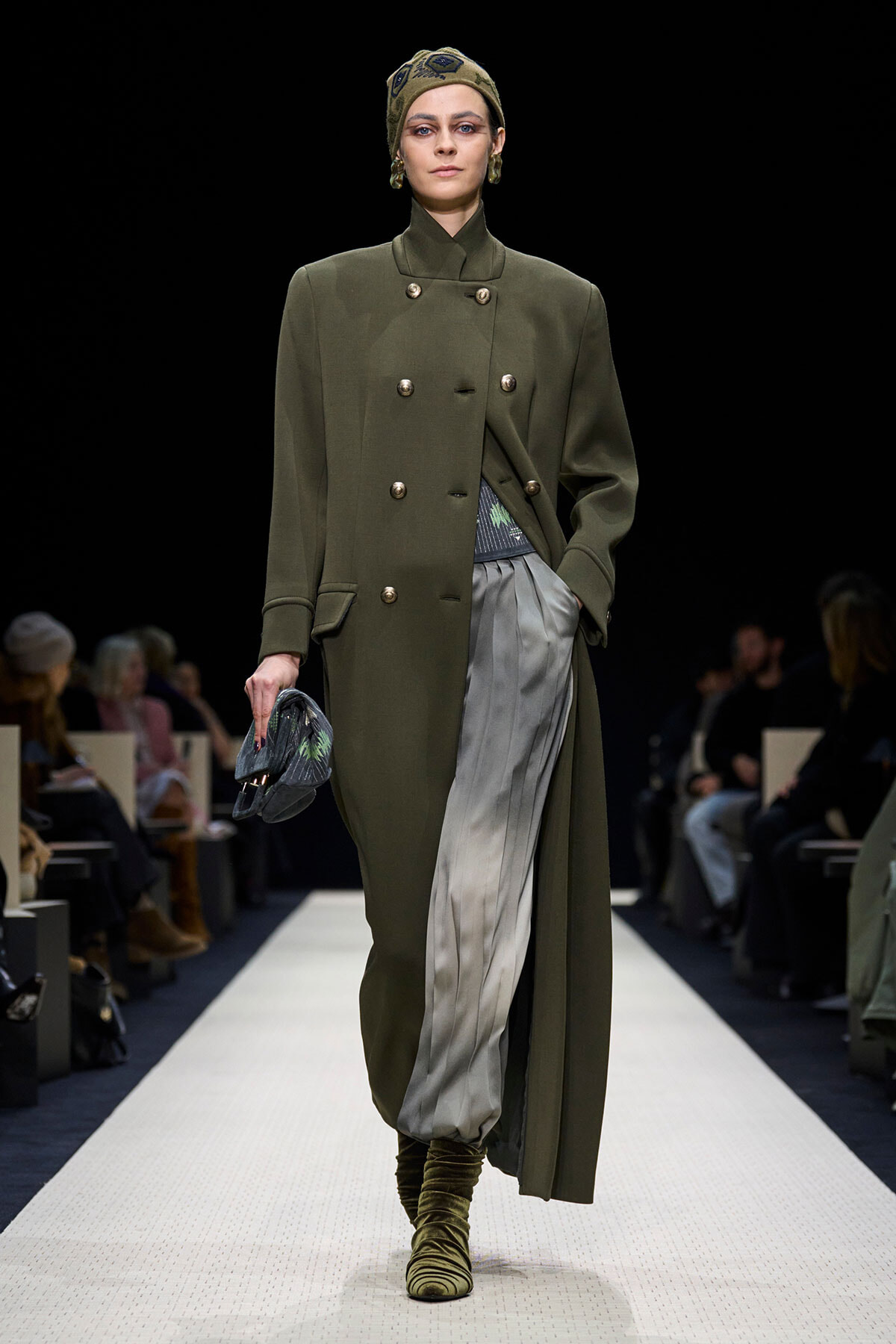 Model walking a runway in an olive double-breasted coat, metallic gray pleated pants, olive boots, and a patterned headscarf with a small clutch in hand.