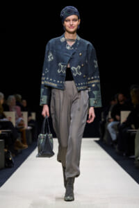 Runway model wearing a dark blue patterned cropped jacket, grey trousers, lace-up boots, and carrying a dark handbag.