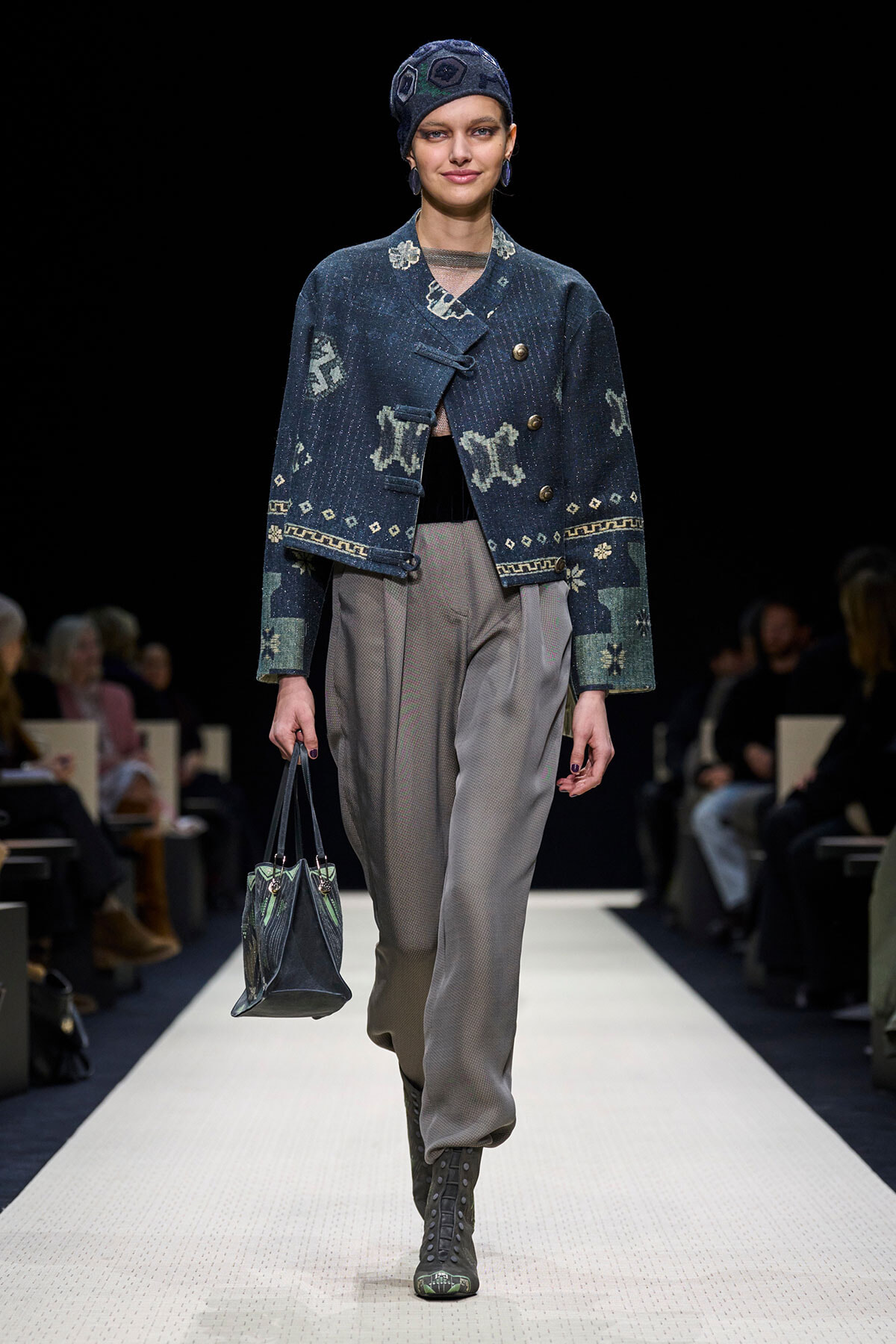 Runway model wearing a dark blue patterned cropped jacket, grey trousers, lace-up boots, and carrying a dark handbag.