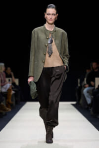 Model walks a runway wearing an olive green blazer over a beige knit top, black satin loose pants, lace-up boots, and holds a green hat.
