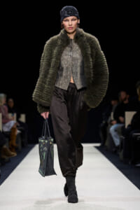 Model on the runway wearing an oversized olive fur coat over a metallic top, black trousers, and black boots, carrying a patterned tote bag.