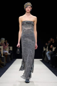 Model walks a fashion runway in a strapless silver mesh gown, with a patterned headwrap and small black handbag.