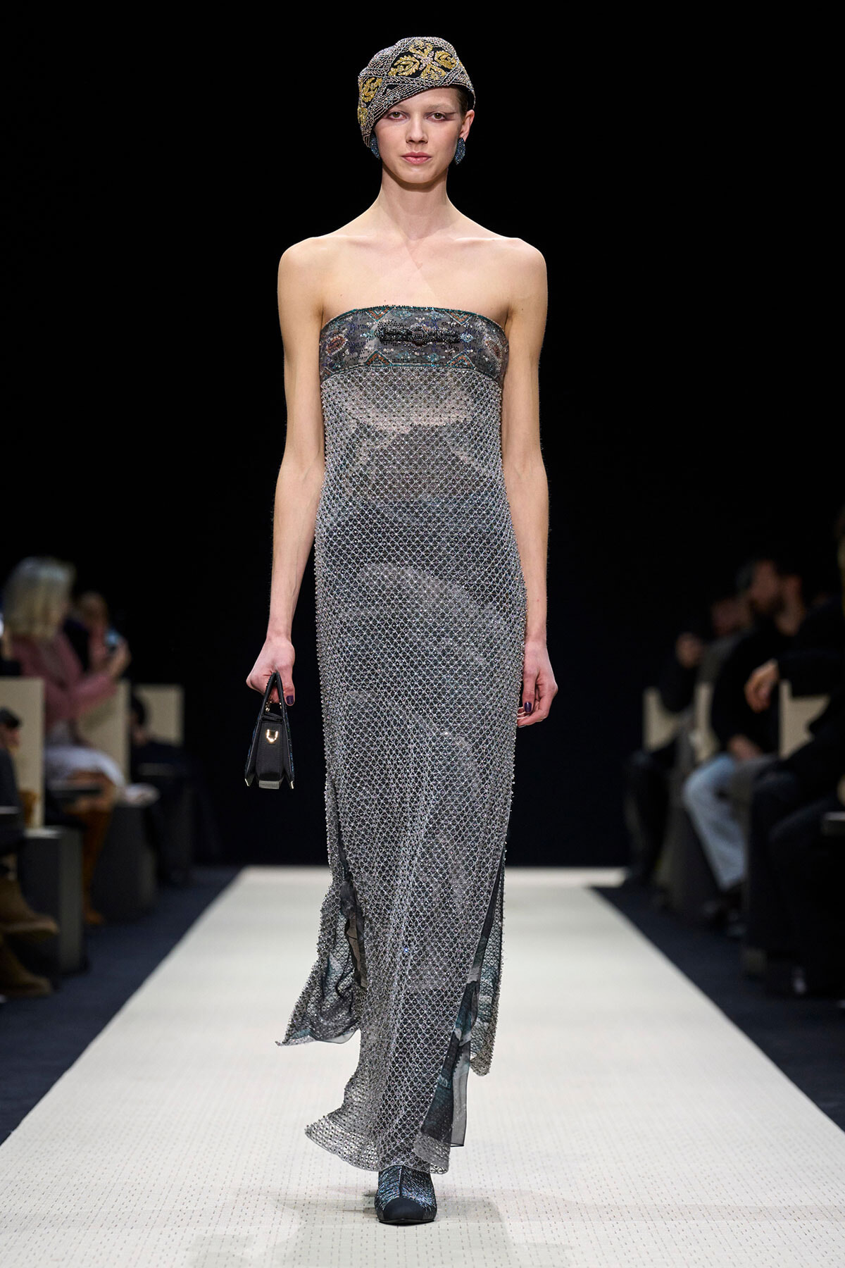 Model walks a fashion runway in a strapless silver mesh gown, with a patterned headwrap and small black handbag.