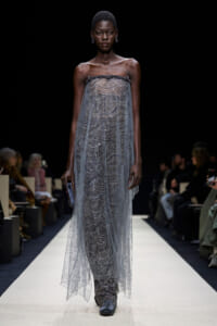 Female model walks a fashion show wearing a floor-length sheer silver beaded gown with a strapless neckline, paired with embellished boots.