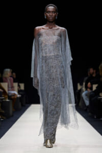 Model walking a runway in a silver, off-shoulder gown with an intricate swirling beaded pattern and a sheer, dotted cape.