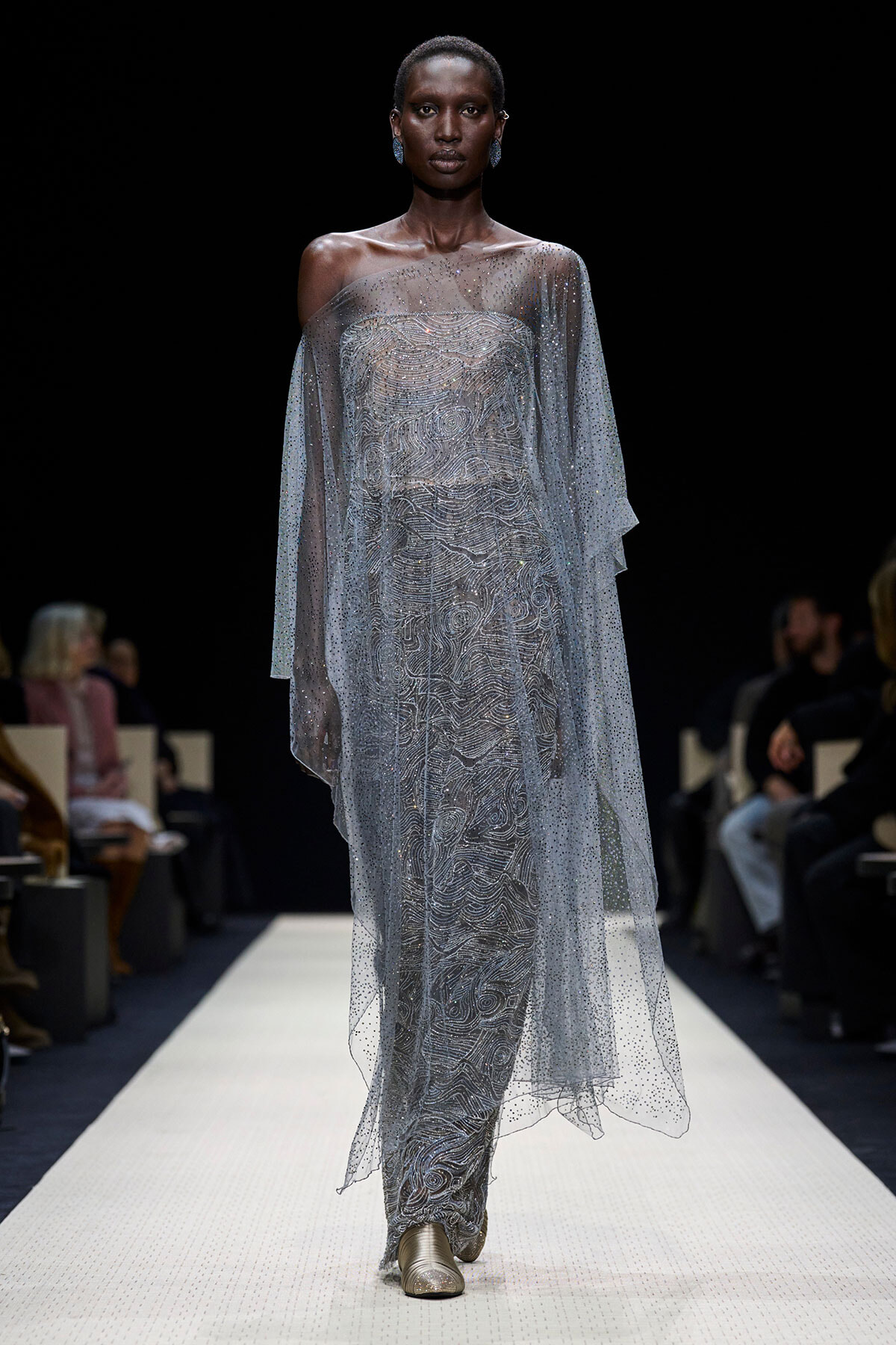 Model walking a runway in a silver, off-shoulder gown with an intricate swirling beaded pattern and a sheer, dotted cape.