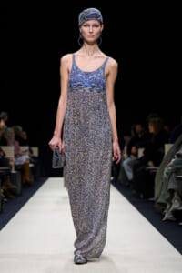 Runway model walking the catwalk in a blue patterned maxi dress with a matching headscarf and large hoop earrings.