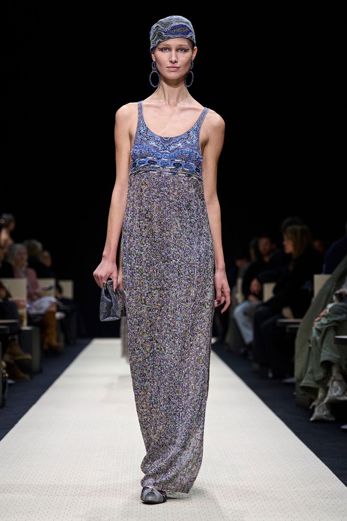 Runway model walking the catwalk in a blue patterned maxi dress with a matching headscarf and large hoop earrings.