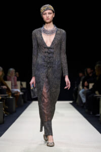 Model walking a metallic beaded gown with deep V neckline and side-slit on a runway, wearing a patterned headscarf and chunky metallic necklace.