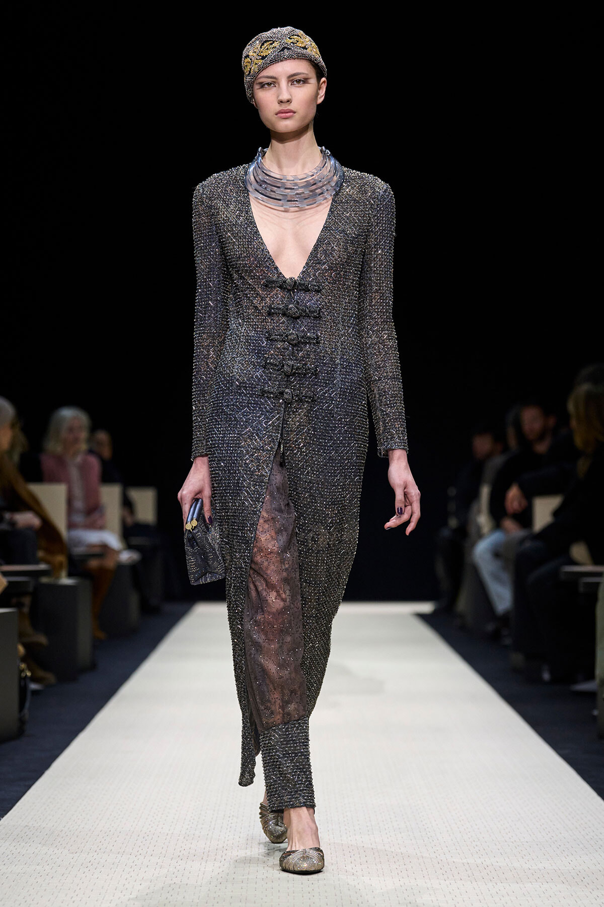 Model walking a metallic beaded gown with deep V neckline and side-slit on a runway, wearing a patterned headscarf and chunky metallic necklace.