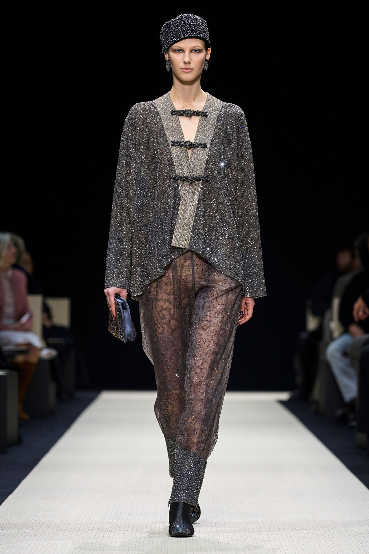 Model on a runway wearing a glittery silver cape-like top with three toggle closures and sheer sequined pants, paired with a matching cap and boots.