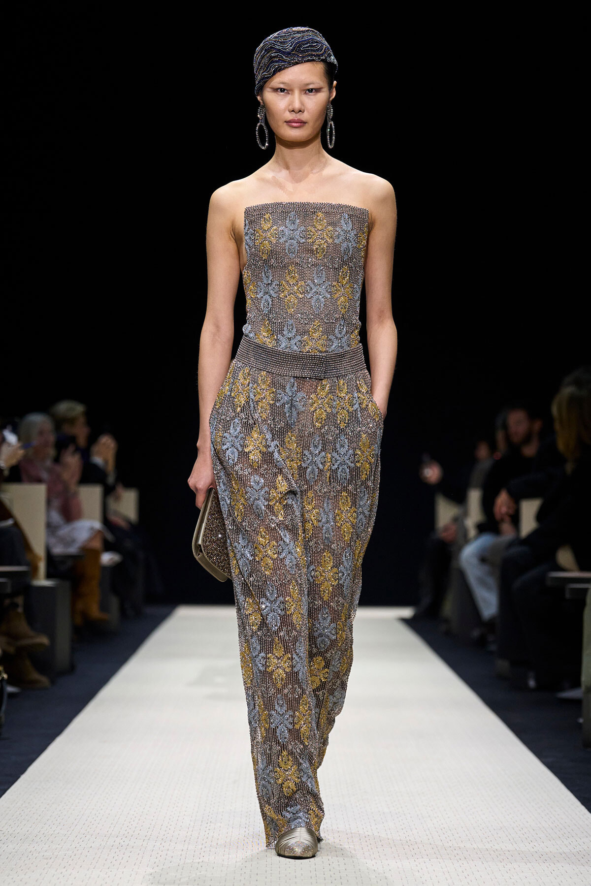 Runway model wearing a sleeveless beaded, gold- and silver-pattern jumpsuit with a matching headwrap and large hoop earrings, carrying a small clutch on the catwalk.