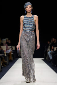 Model walking a runway in a blue-green beaded sleeveless top and a silver, sequined, floor-length skirt with a matching head wrap and dangling blue earrings, holding a small clutch.