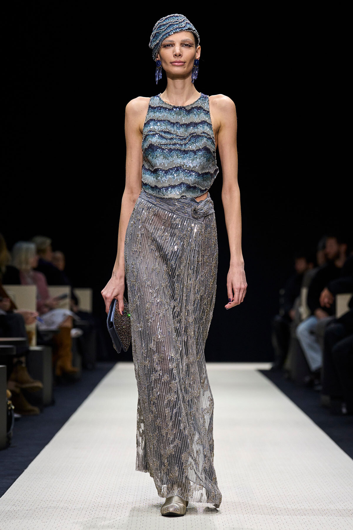 Model walking a runway in a blue-green beaded sleeveless top and a silver, sequined, floor-length skirt with a matching head wrap and dangling blue earrings, holding a small clutch.