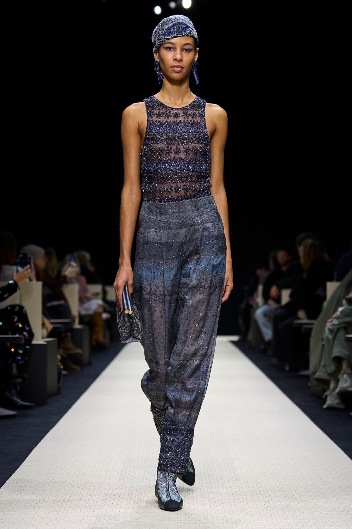 Runway model wearing a sheer beaded blue top and glittery gray pants, with a blue patterned headwrap and blue earrings, holding a small clutch.