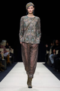 Model walks a fashion runway wearing a metallic beaded jacket, sheer jeweled pants, a patterned headwrap, and large hoop earrings.