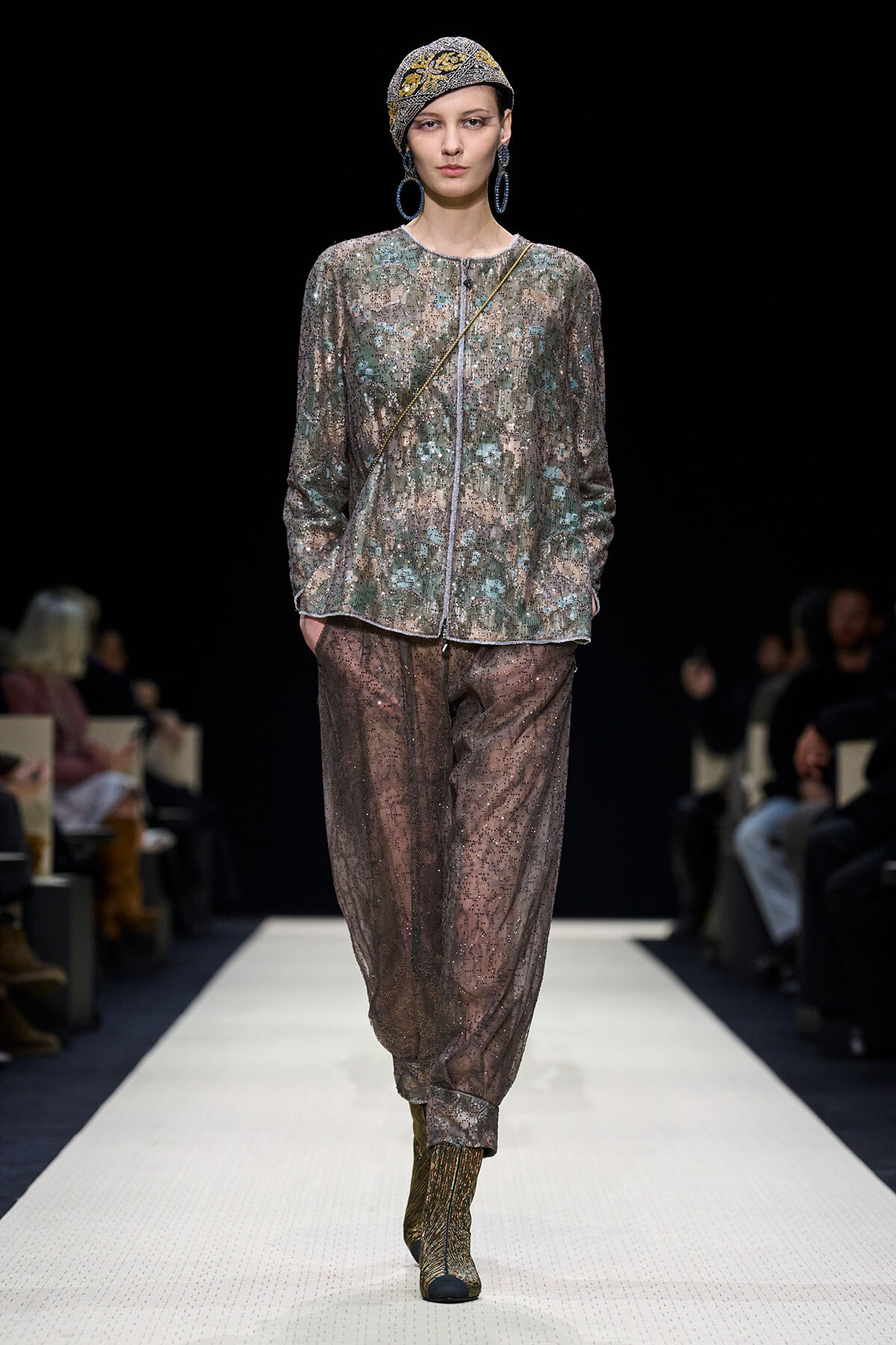 Model walks a fashion runway wearing a metallic beaded jacket, sheer jeweled pants, a patterned headwrap, and large hoop earrings.