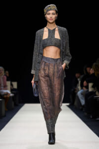 Model on a runway wearing a glittery silver ensemble: cropped top, matching jacket, and sheer beaded pants with a headwrap.