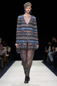 Model walking a runway in a blue-brown embroidered coat over sheer lace pants, with a jeweled headwrap and blue earrings, clutch in hand.