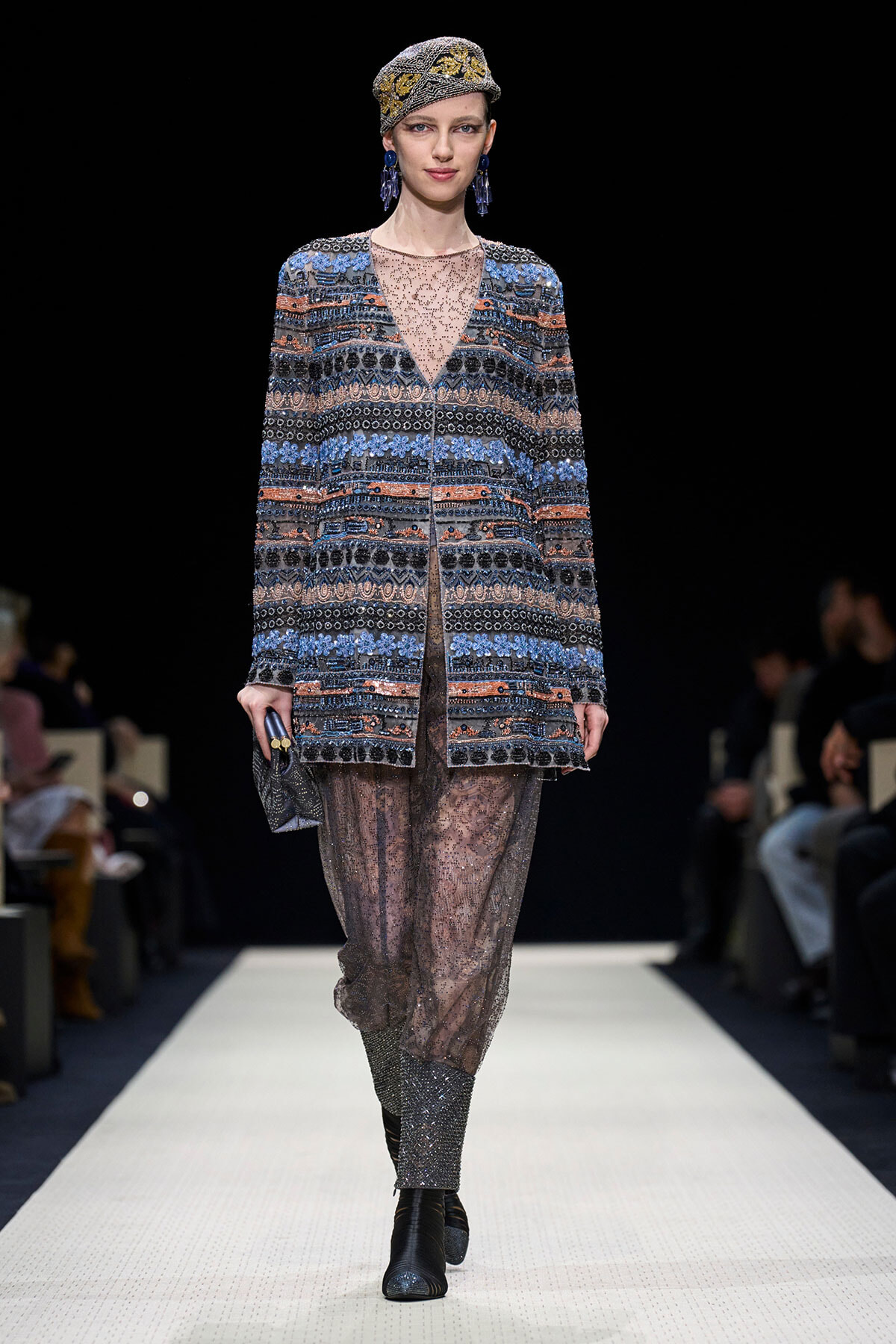 Model walking a runway in a blue-brown embroidered coat over sheer lace pants, with a jeweled headwrap and blue earrings, clutch in hand.