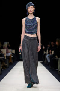 Model walks a runway in a blue-green sequined halter top with a glittery gray wrap skirt and teal shoes, accessorized with a turban and blue earrings.