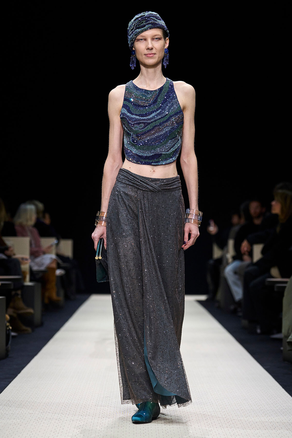 Model walks a runway in a blue-green sequined halter top with a glittery gray wrap skirt and teal shoes, accessorized with a turban and blue earrings.