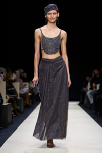 Model walks a fashion runway wearing a glittering silver cropped top and matching long skirt, with a wrapped headscarf and large hoop earrings, carrying a small clutch.