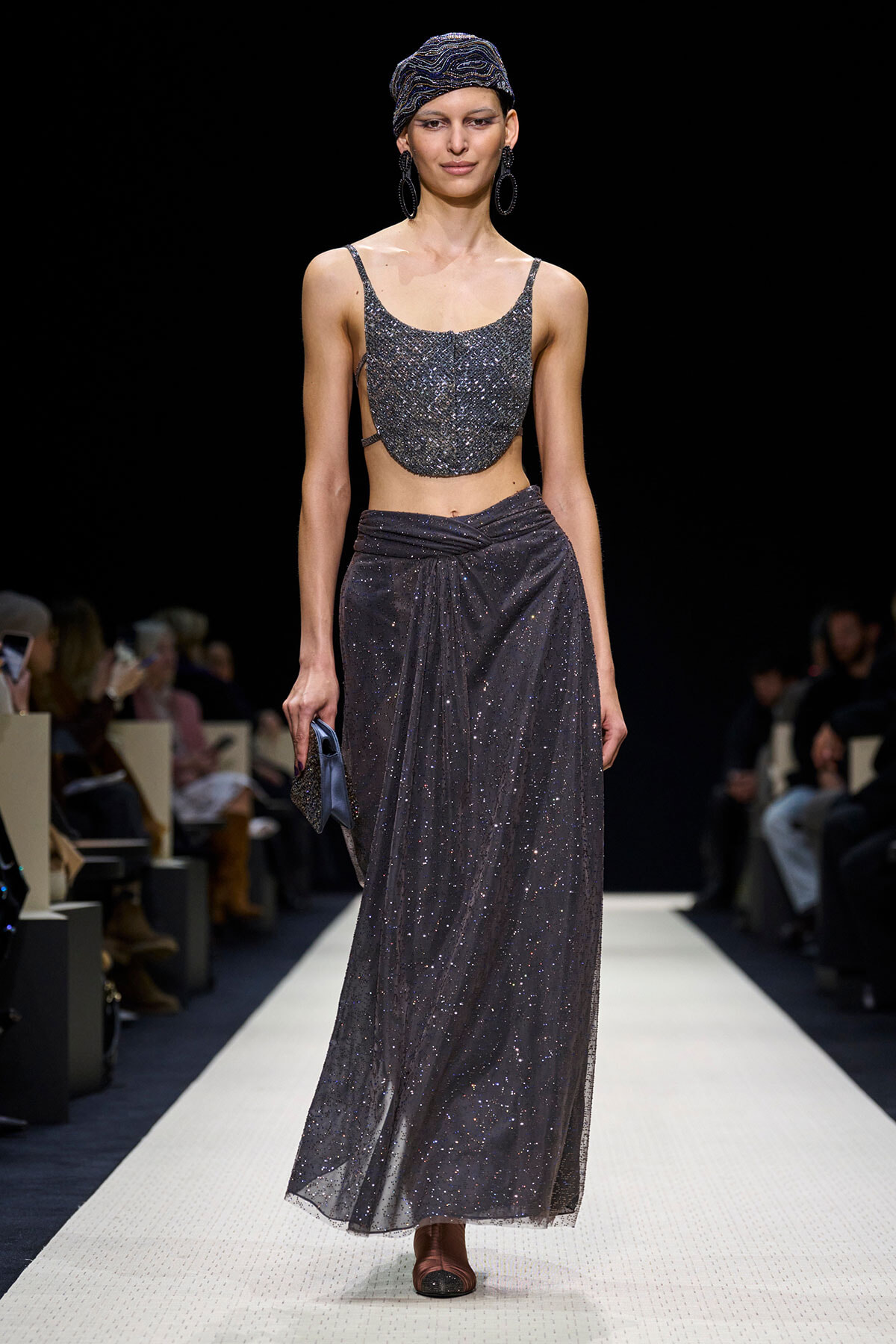 Model walks a fashion runway wearing a glittering silver cropped top and matching long skirt, with a wrapped headscarf and large hoop earrings, carrying a small clutch.