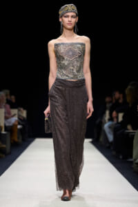 Model wearing a jeweled strapless top and glittery wide-leg pants on a runway, with a patterned headscarf and dangling earrings.