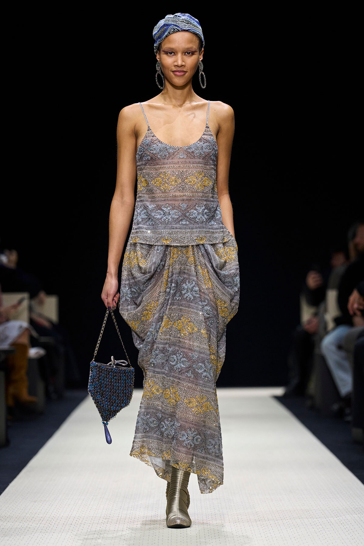 Model on a runway wearing a silver patterned dress with gold accents, blue headwrap, large hoop earrings, and a blue beaded handbag.