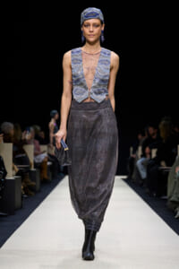Model walks a white runway in a metallic blue-gray ensemble: sleeveless jeweled mesh top with matching vest panels, wide harem-style sequin pants, a matching head wrap, blue earrings, and a small clutch purse in hand.